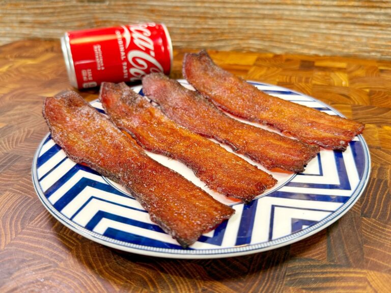 Four strips of crispy, glazed bacon on a blue-and-white zigzag patterned plate, with a partially opened can of Coca-Cola in the background, set on a wooden surface.