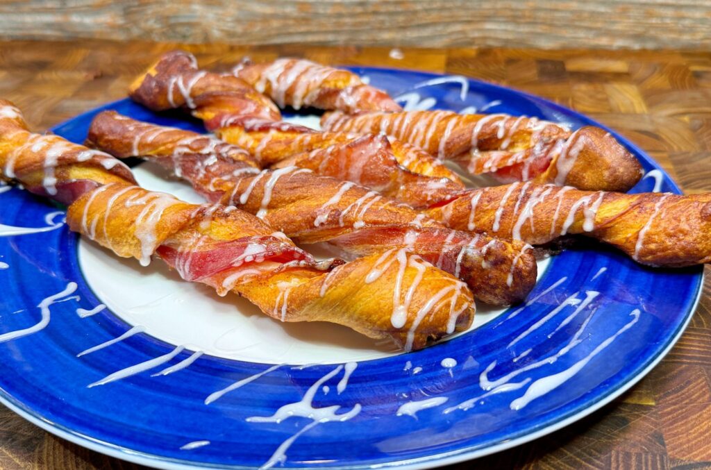 Twisted pastry sticks drizzled with icing are arranged on a blue plate with a white center. The wooden background adds a rustic touch to the presentation.