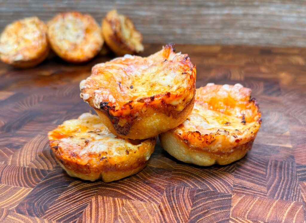 Three mini deep-dish pizzas stacked on a wooden surface, with a crispy, golden-brown crust and melted cheese on top. More pizzas are blurred in the background.