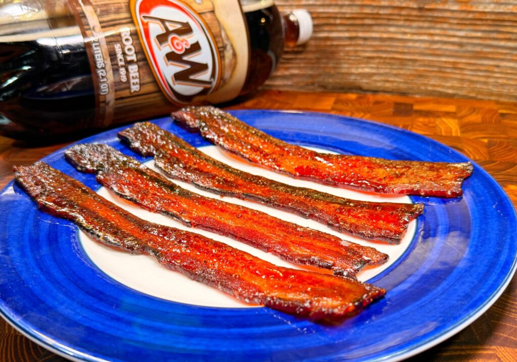 Four strips of candied bacon are neatly arranged on a blue and white plate. In the background, theres a bottle of A&W root beer lying on its side on a wooden surface.