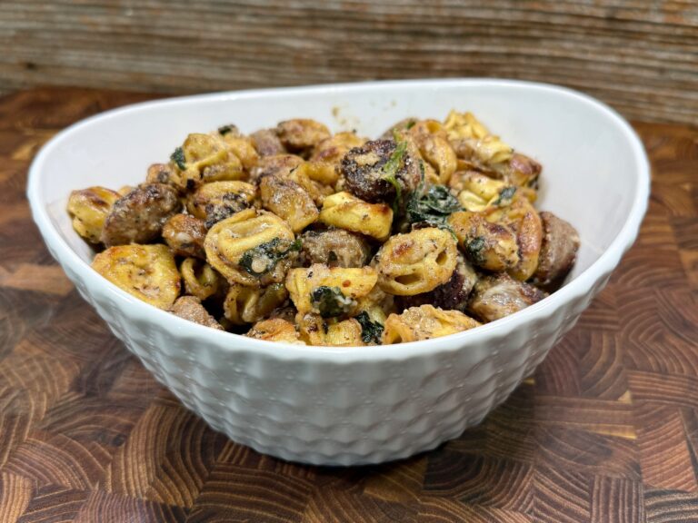 A white textured bowl filled with cooked tortellini pasta, sausage slices, and leafy greens sits on a wooden surface with a rustic wood background.