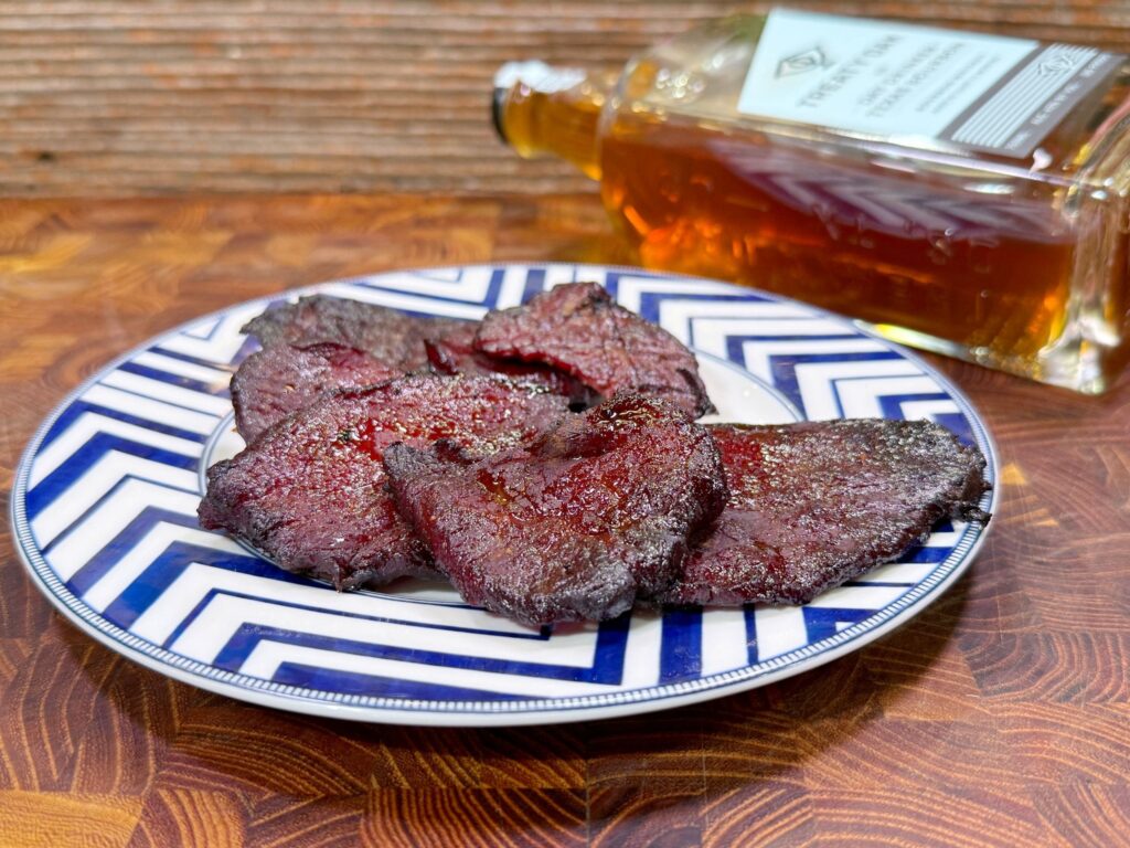 A plate with several pieces of dark, seasoned beef jerky sits on a blue and white patterned dish. In the background, a bottle of whiskey lies on its side on a wooden surface.