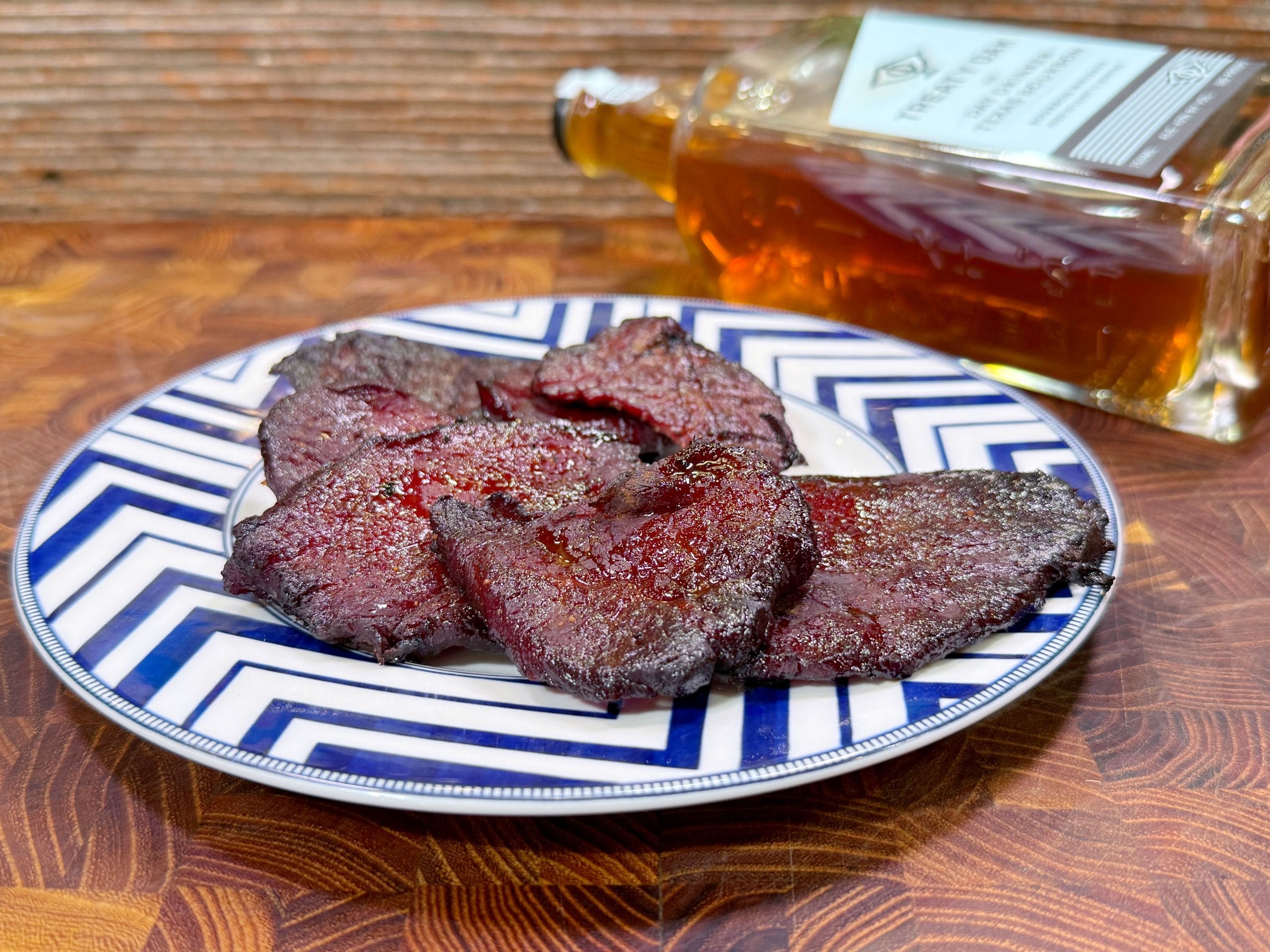 A plate with several pieces of dark, seasoned beef jerky sits on a blue and white patterned dish. In the background, a bottle of whiskey lies on its side on a wooden surface.