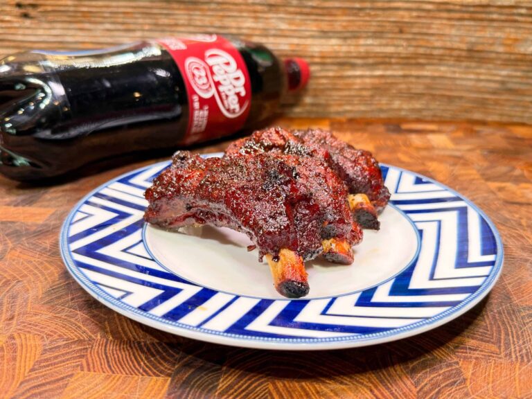 A plate with grilled ribs sits on a blue and white zigzag-patterned dish, with a bottle of Dr Pepper soda lying on its side in the background on a wooden surface.