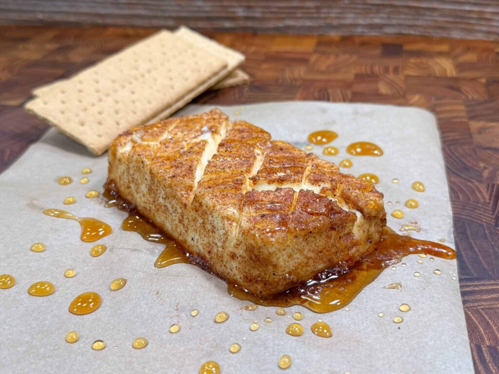 A block of seasoned, grilled cream cheese with a caramelized glaze, drizzled with honey and served on parchment paper, with two graham crackers in the background.