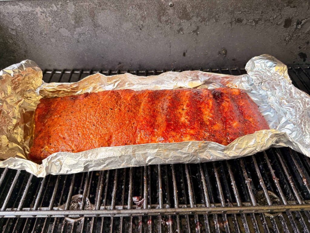 A rack of seasoned ribs covered in foil is cooking on a grill, with visible grill grates beneath and smoke marks on the meat.