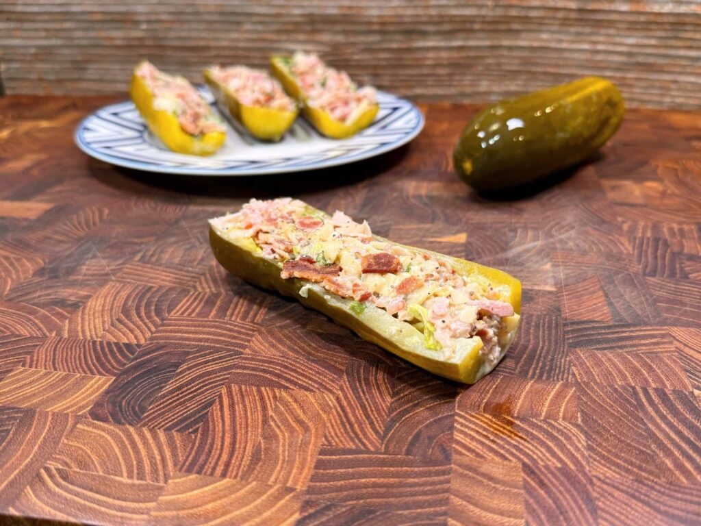 A pickle half stuffed with a creamy mixture sits on a wooden surface, with a whole pickle and a plate of more stuffed pickle halves in the background.