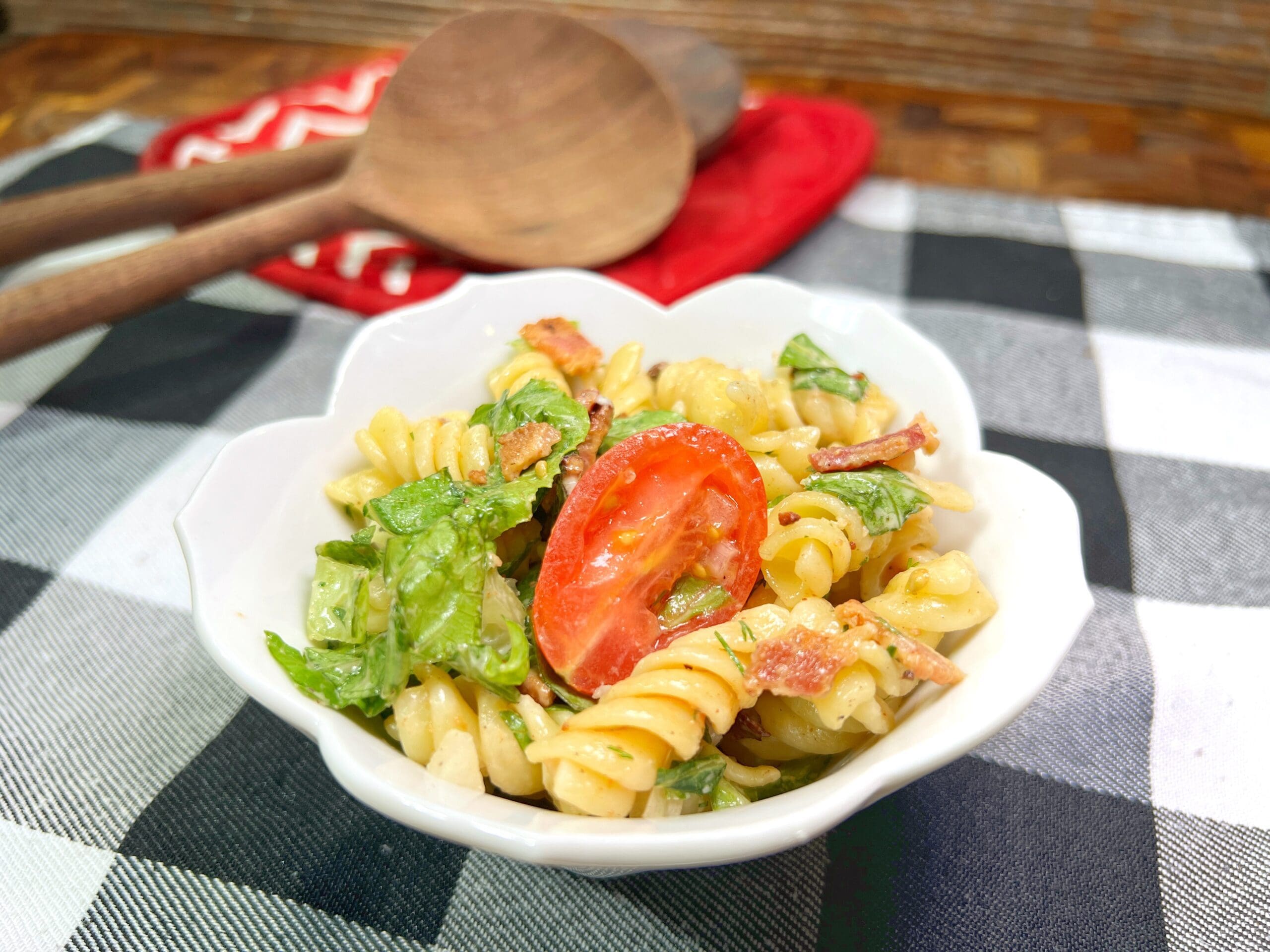 A white bowl filled with rotini pasta salad, mixed with lettuce, bacon pieces, and a slice of tomato, sits on a black-and-white checkered cloth near wooden salad utensils and a red pot holder.