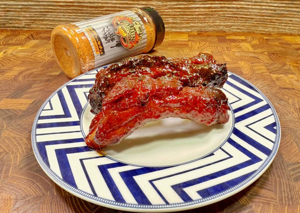 A plate with two glazed, barbecued ribs sits on a blue and white zigzag-patterned plate. Behind the plate is a bottle of barbecue seasoning tipped on its side on a wooden surface.