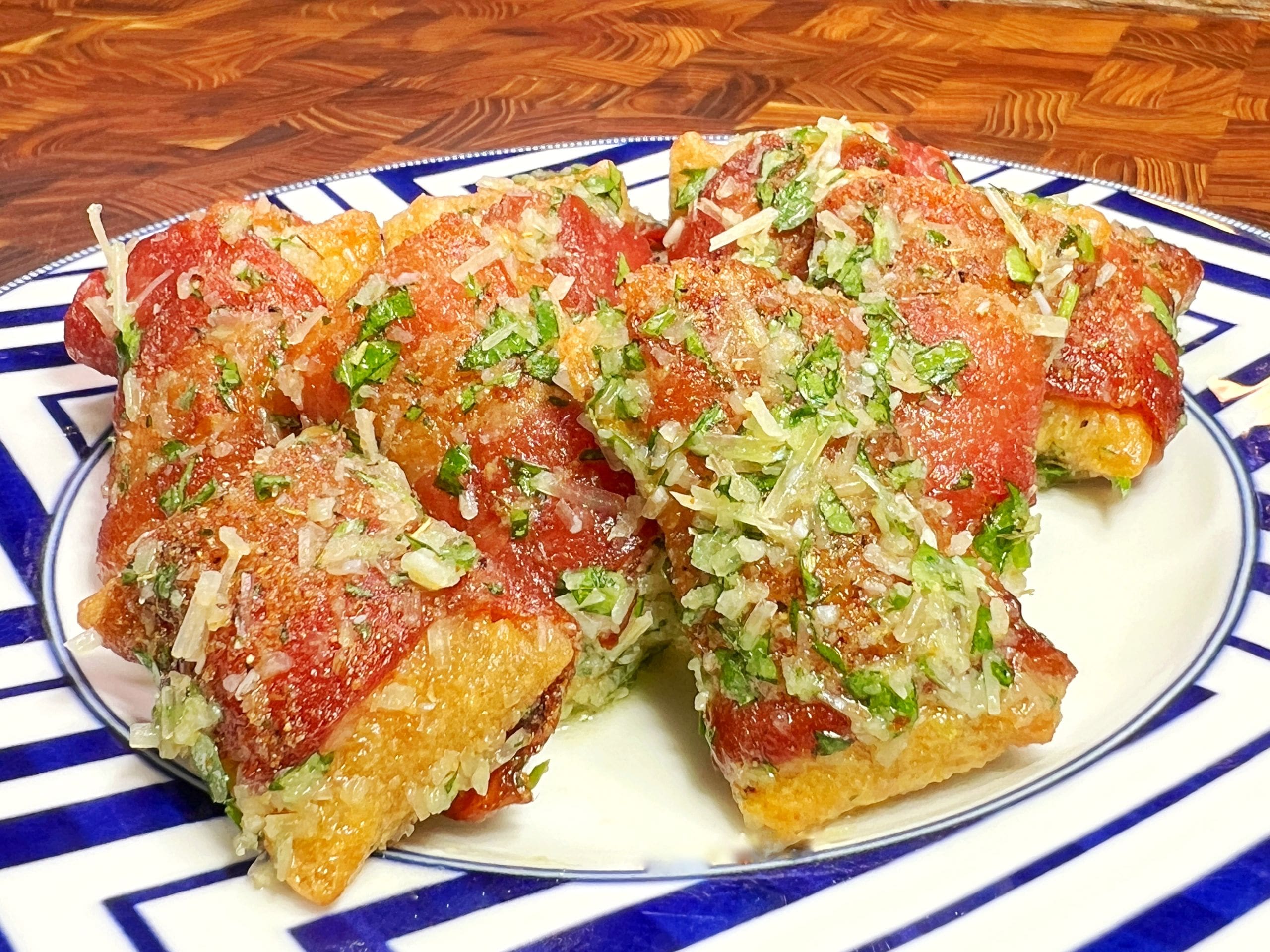 Rectangular slices of Detroit-style pizza are topped with tomato sauce, grated cheese, and chopped herbs, arranged on a white plate with a blue geometric border on a wooden surface.