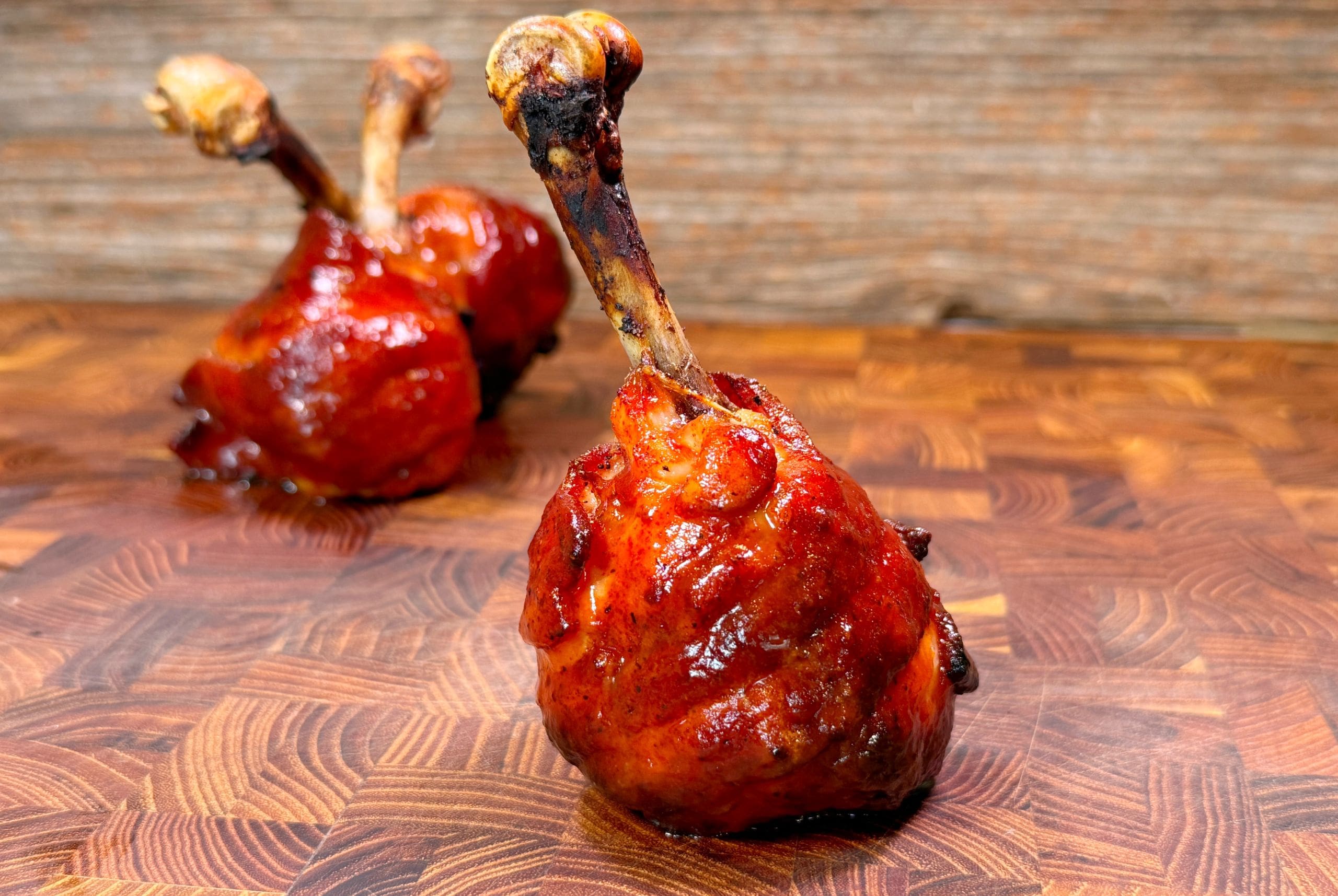 Two chicken drumsticks are cooked and presented upright on a wooden surface, with crispy, glazed skin and exposed clean bones. The background is a wooden texture, slightly blurred.