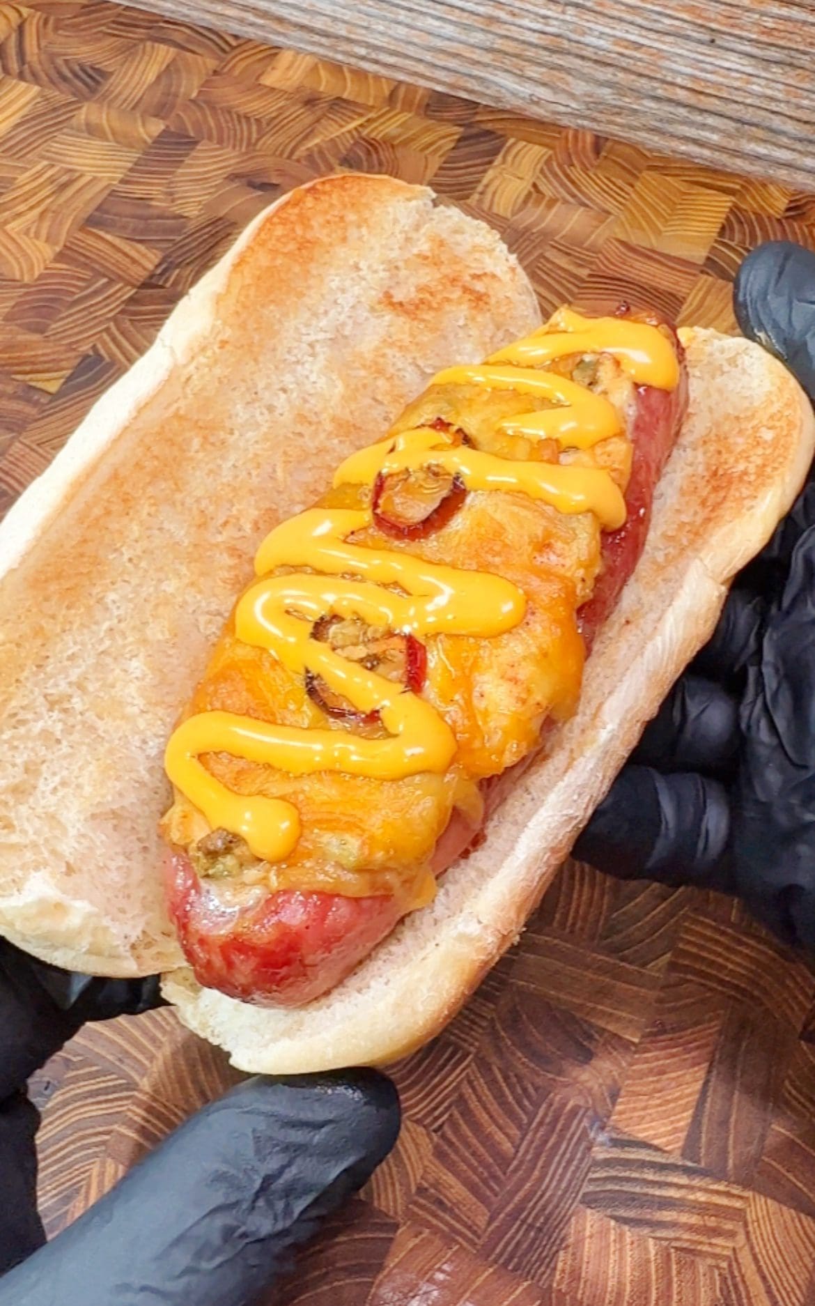 A hot dog in a bun topped with melted cheese, grilled onions, and drizzled yellow sauce, held by hands wearing black gloves over a wooden surface.