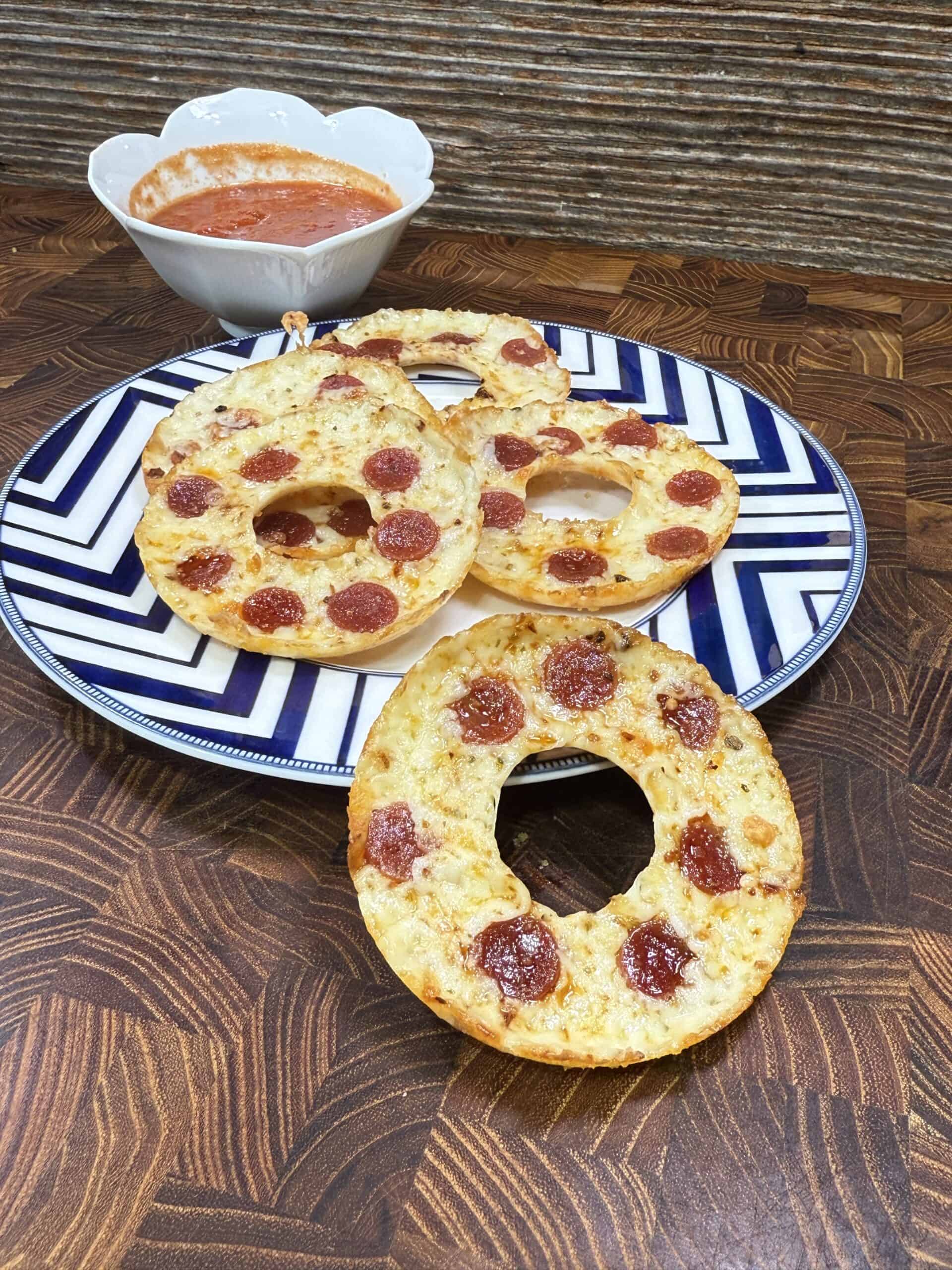 Three bagel halves topped with melted cheese and pepperoni slices are arranged on a blue and white plate, inspired by the Best Pepperoni Pizza Rings Recipe, with a bowl of red dipping sauce in the background on a wooden surface.