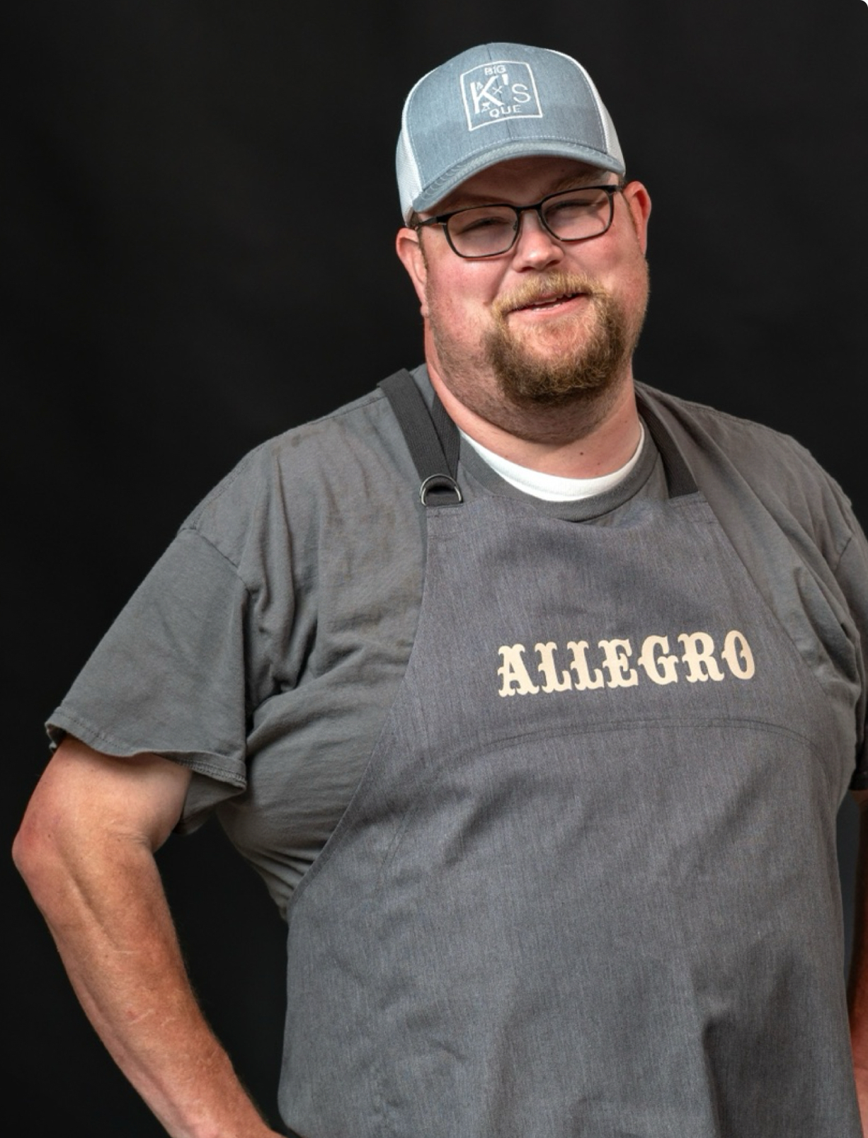 A man wearing glasses, a gray t-shirt, a gray apron labeled ALLEGRO, and a blue baseball cap stands smiling against a black background.