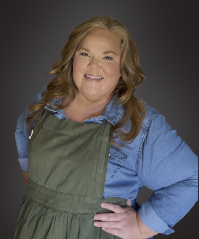 A smiling woman with wavy, light brown hair wears a green apron over a blue denim shirt, standing with one hand on her hip against a dark background.