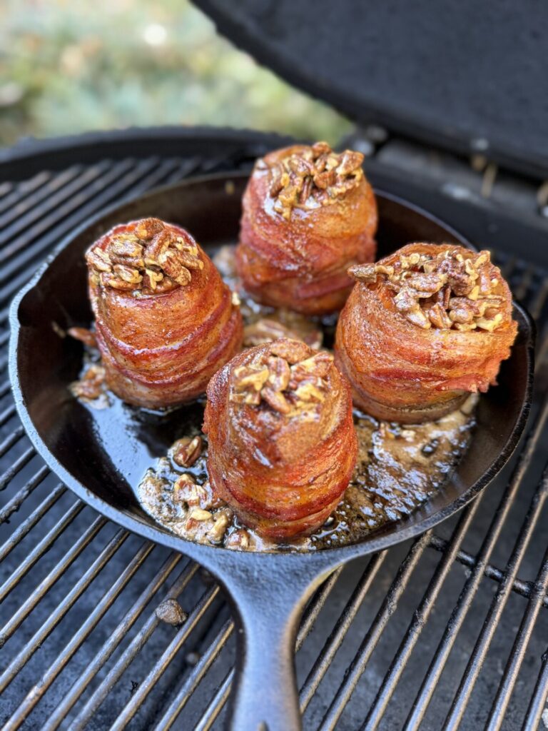 Four bacon-wrapped, pecan-topped meat bundles cook in a cast iron skillet on a grill, with juices sizzling around them. The background is blurred with hints of greenery.