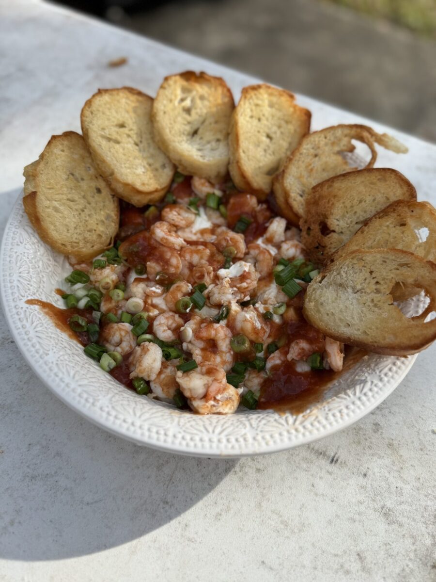 A white bowl filled with a shrimp and tomato mixture, garnished with chopped green onions, is surrounded by toasted slices of bread, arranged in a fan shape on the edge of the bowl, sitting on a white surface.
