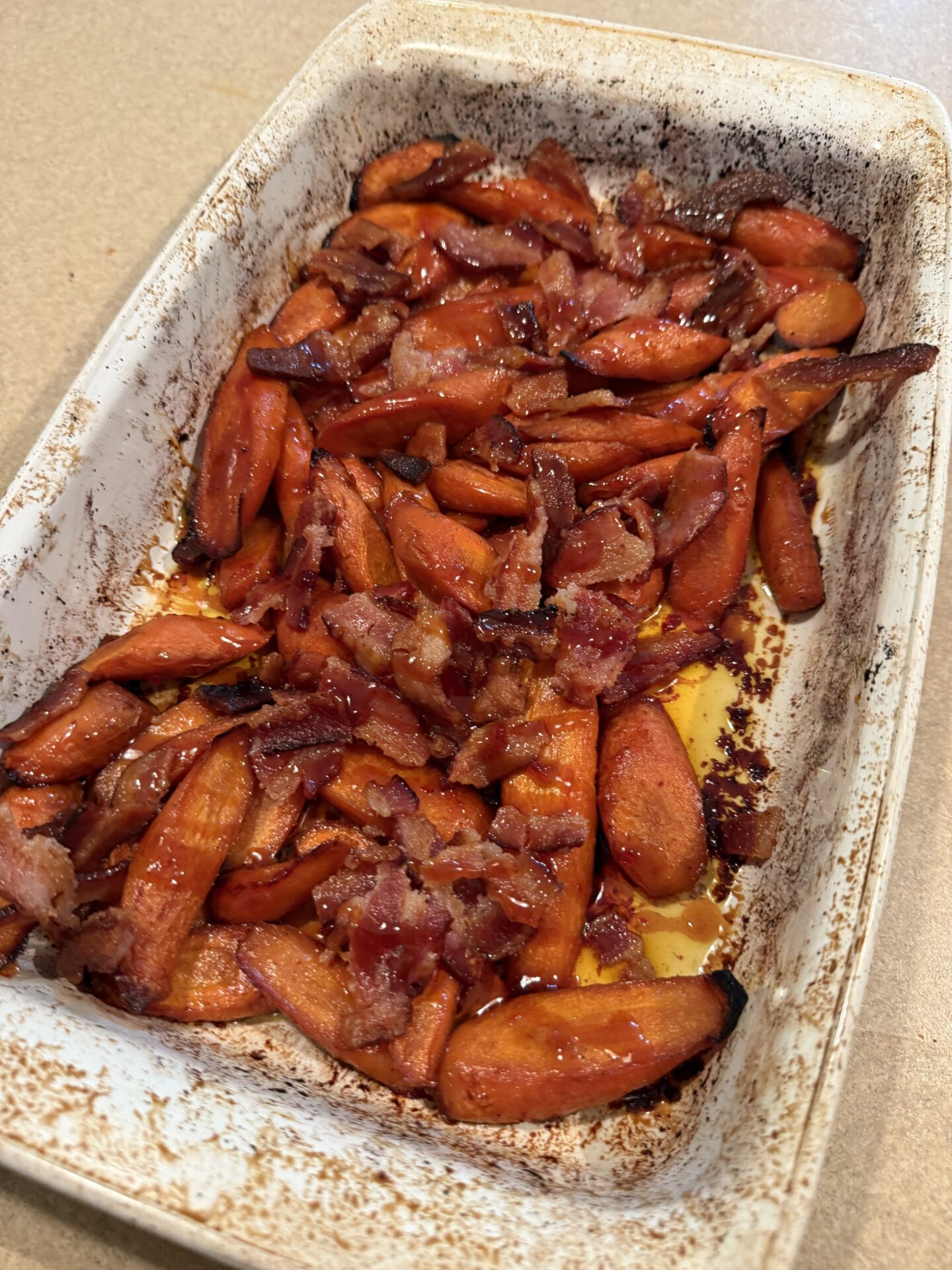 A white baking dish holds roasted carrot slices topped with crispy bacon pieces. The carrots are slightly caramelized, and the dish shows baked-on residue around the edges.