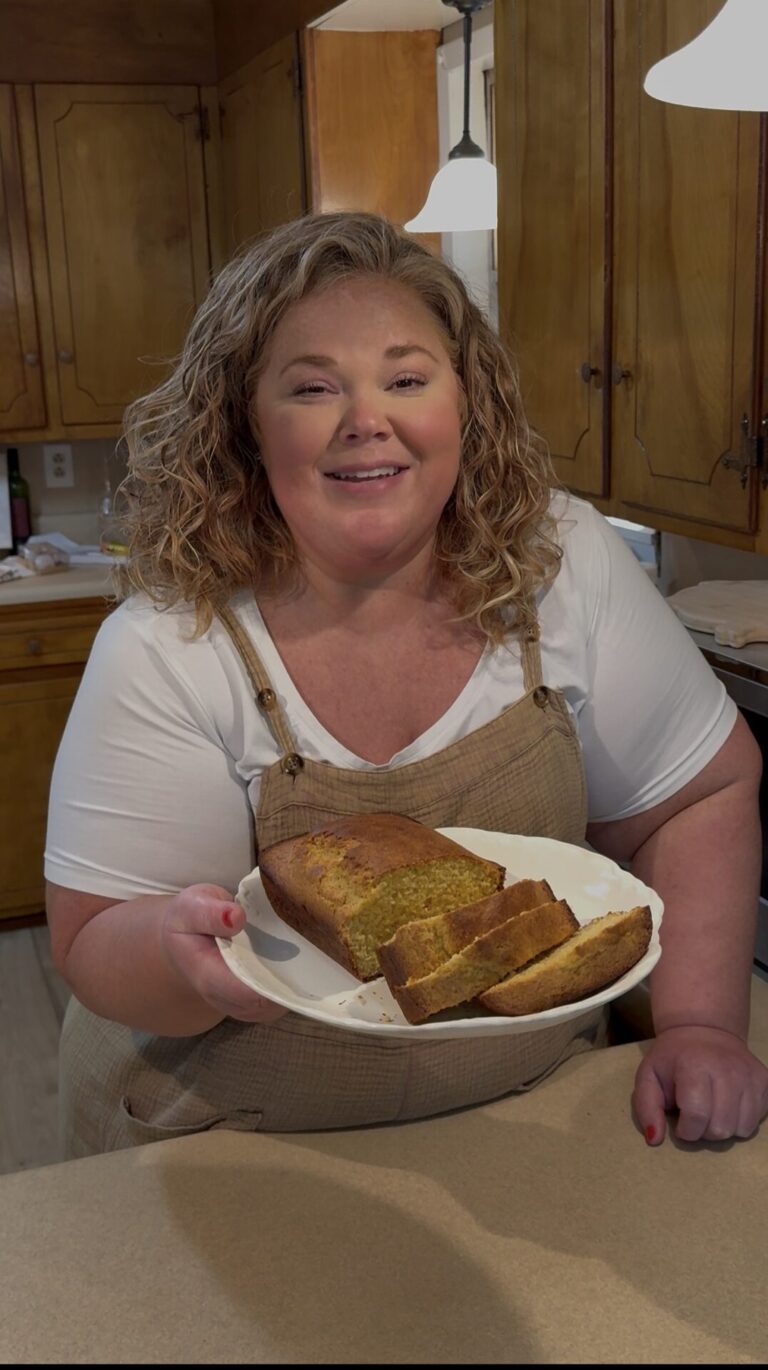 A woman with curly hair, wearing a white shirt and beige apron, smiles in a kitchen while holding a plate with a loaf of sliced bread. Wooden cabinets and kitchen items are visible in the background.