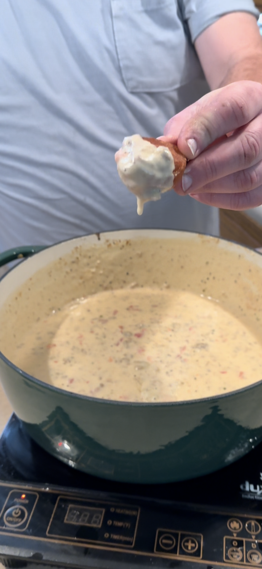 A person holds a piece of bread dipped in creamy soup or sauce over a green pot filled with the same mixture, standing next to a cooktop.