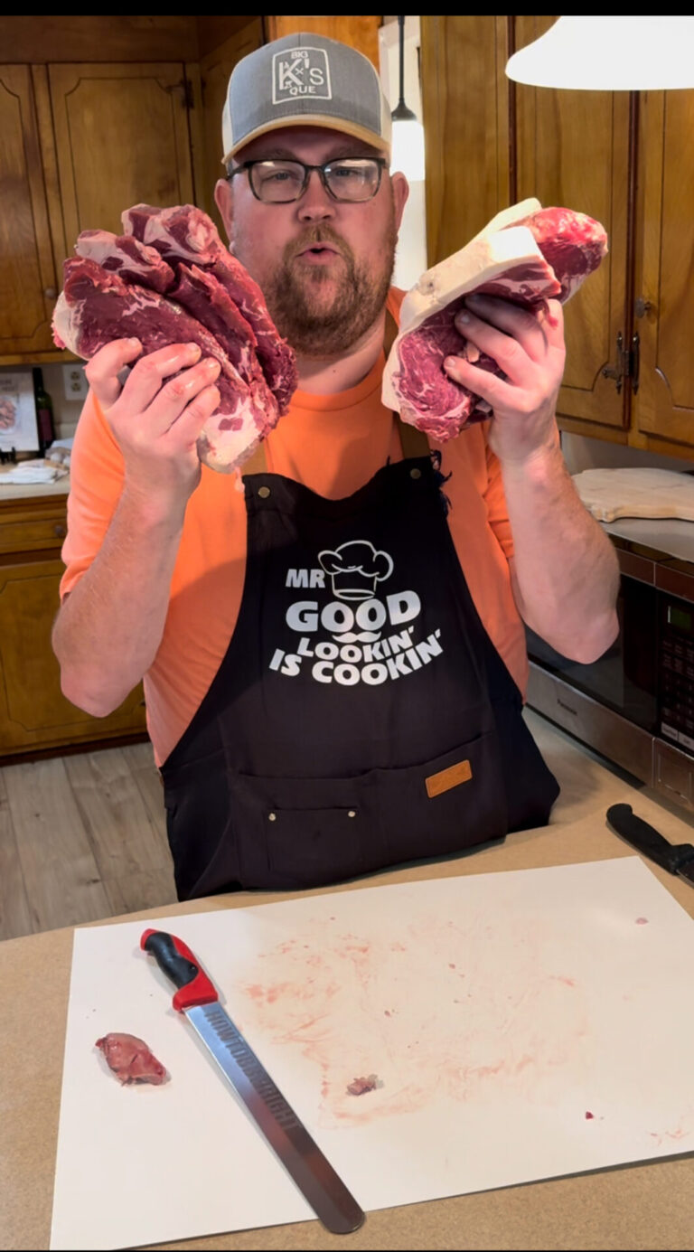 A man in an apron and hat stands in a kitchen, holding up two large raw steaks, one in each hand. A knife and cutting board with meat scraps are on the counter in front of him.