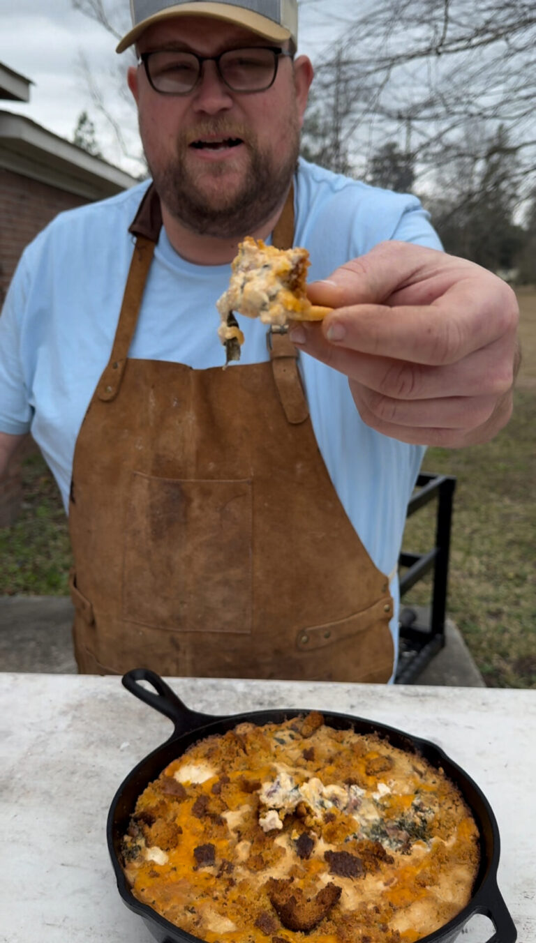A man wearing a brown apron and blue shirt holds out a chip with dip, standing outdoors by a table with a skillet of cheesy dip in front of him.