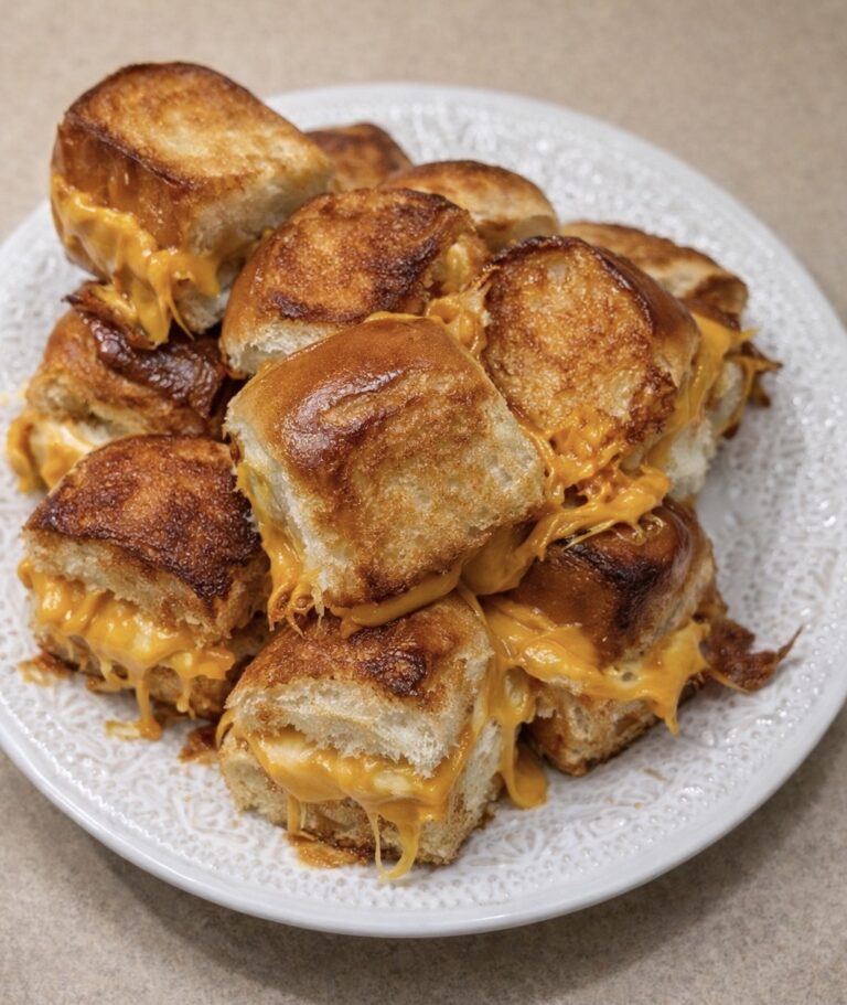 A plate stacked with golden-brown, cheesy slider sandwiches, with melted cheddar cheese oozing between the soft toasted buns. The plate sits on a light-colored countertop.