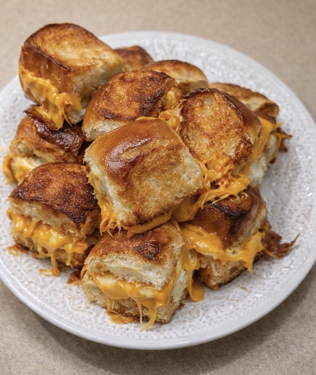 A plate stacked with golden-brown, cheesy slider sandwiches, with melted cheddar cheese oozing between the soft toasted buns. The plate sits on a light-colored countertop.