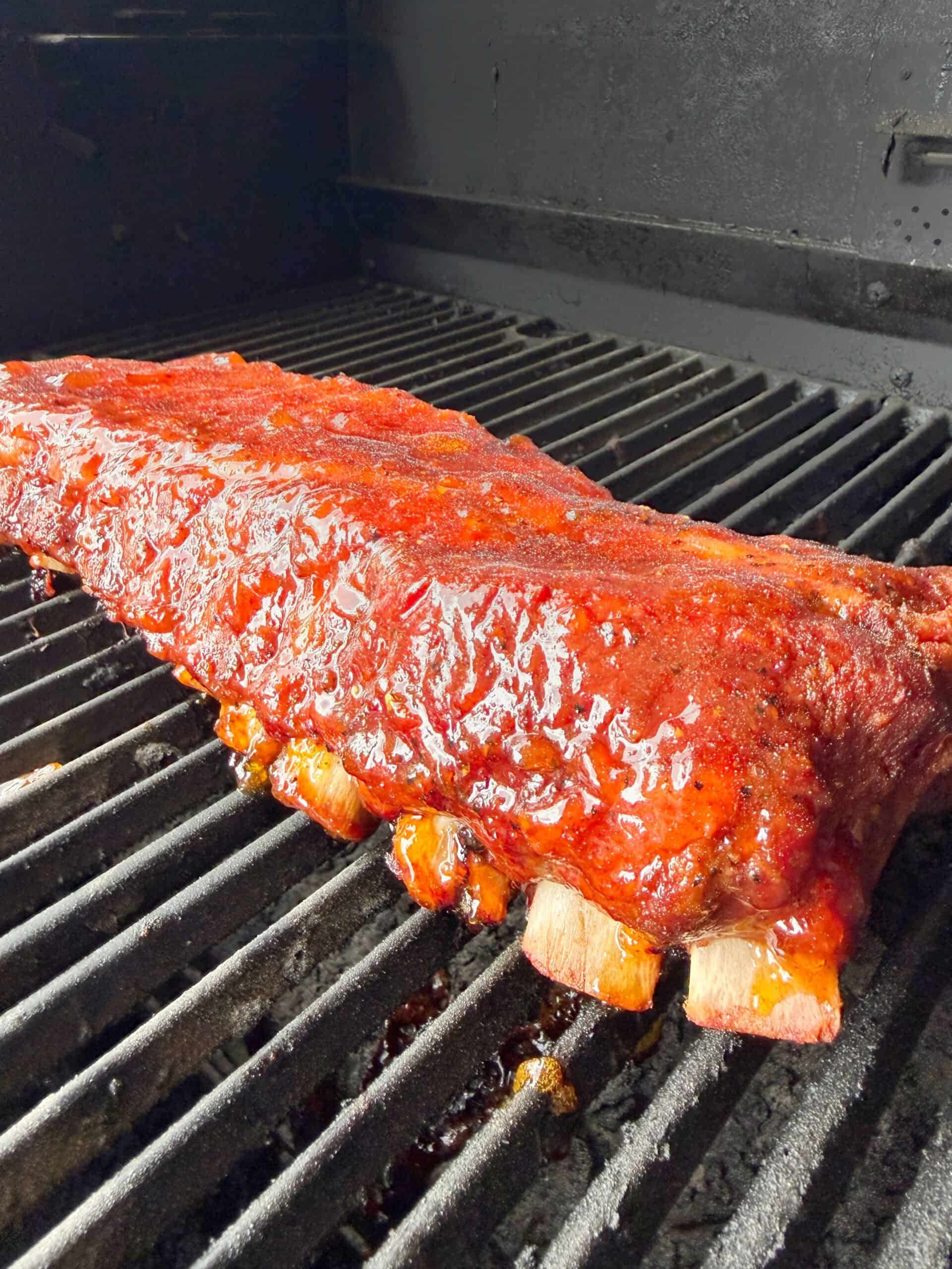A rack of glazed barbecue ribs is cooking on a grill, with the meat glistening and caramelized, and grill marks visible beneath.
