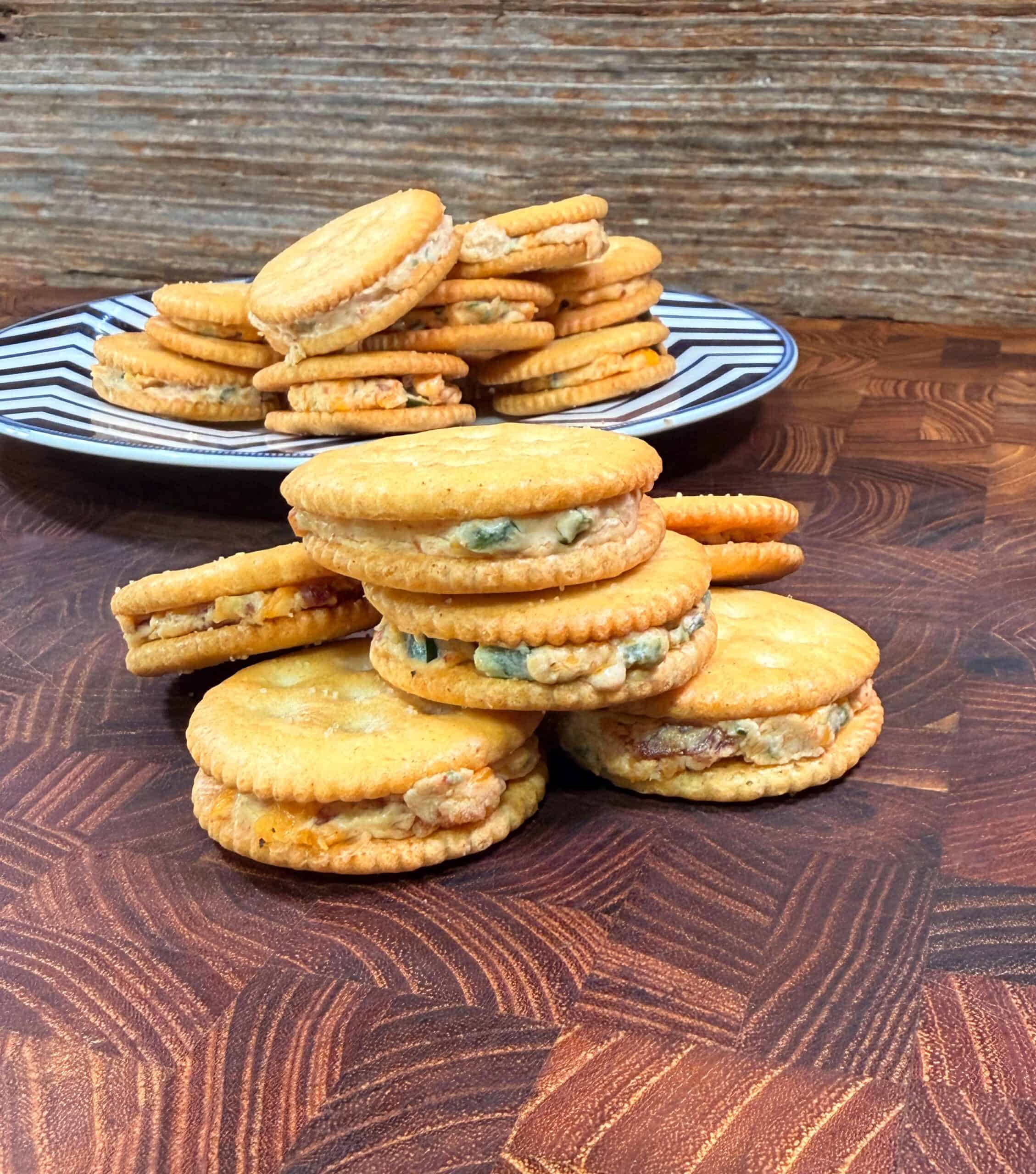 Three stacks of round crackers filled with a creamy mixture are arranged on a wooden surface, with more cracker sandwiches piled on a black-and-white striped plate in the background.
