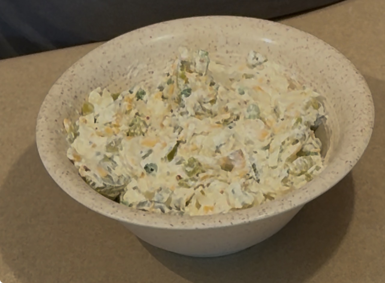 A beige speckled bowl filled with a creamy salad mixture that includes chopped pickles, peas, and shredded cheese, sitting on a light brown countertop.