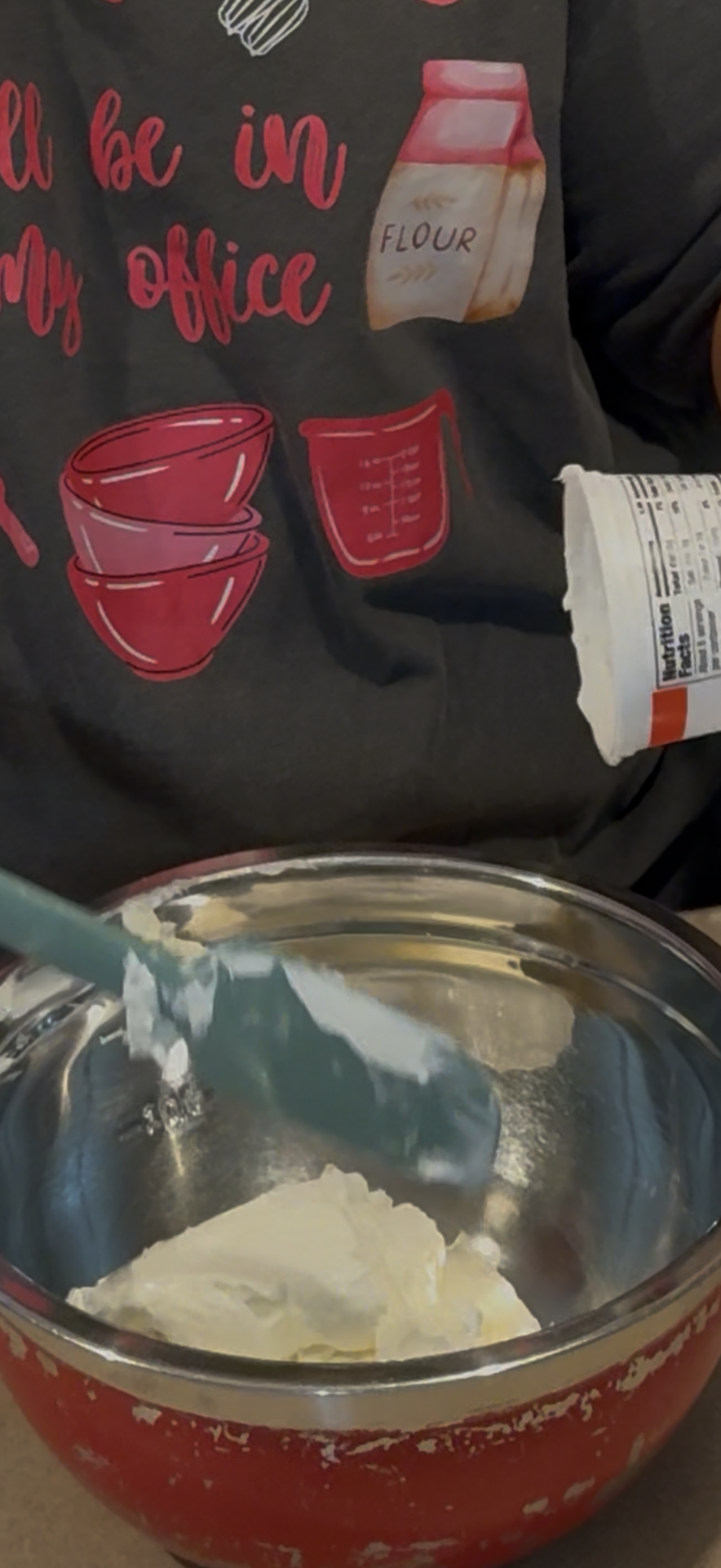 A person in a baking-themed shirt uses a spatula to scoop creamy cheese from a container into a metal mixing bowl.