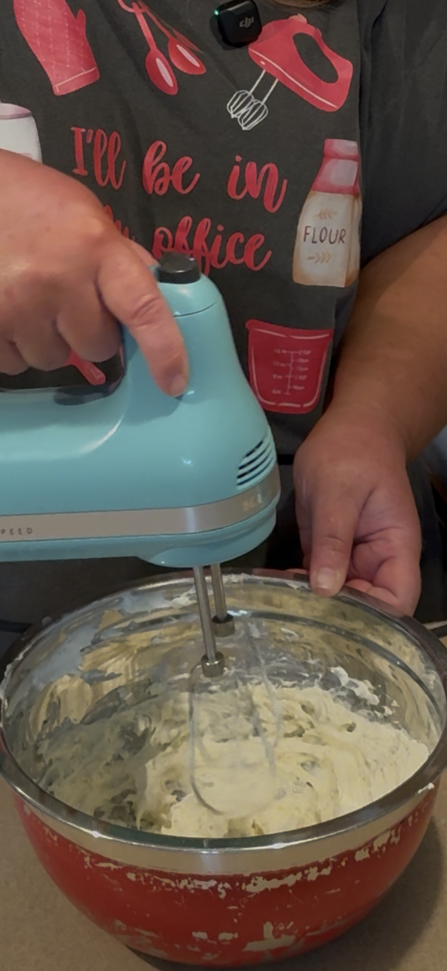A person uses a blue hand mixer to blend ingredients in a red mixing bowl. The person is wearing a dark shirt with baking-themed graphics and text.