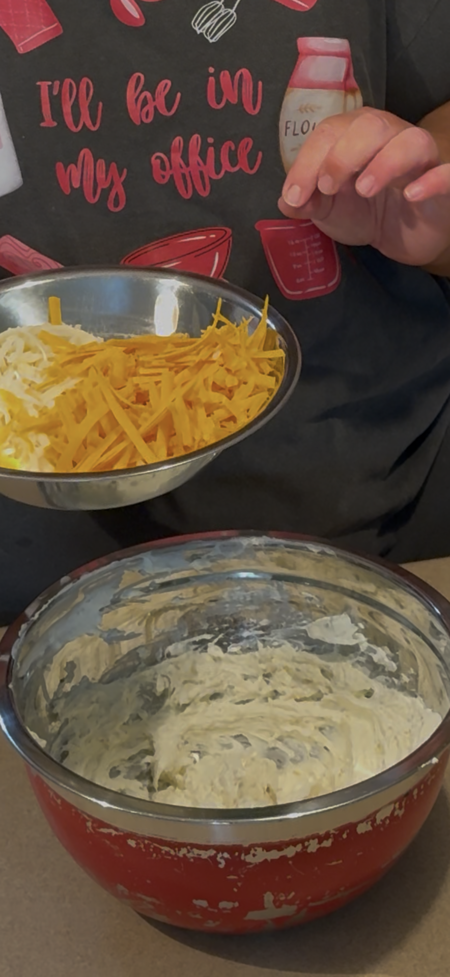 A person holds a metal bowl of shredded cheese above a red mixing bowl filled with creamy batter, preparing to add the cheese. The person is wearing a black shirt with baking-themed graphics.
