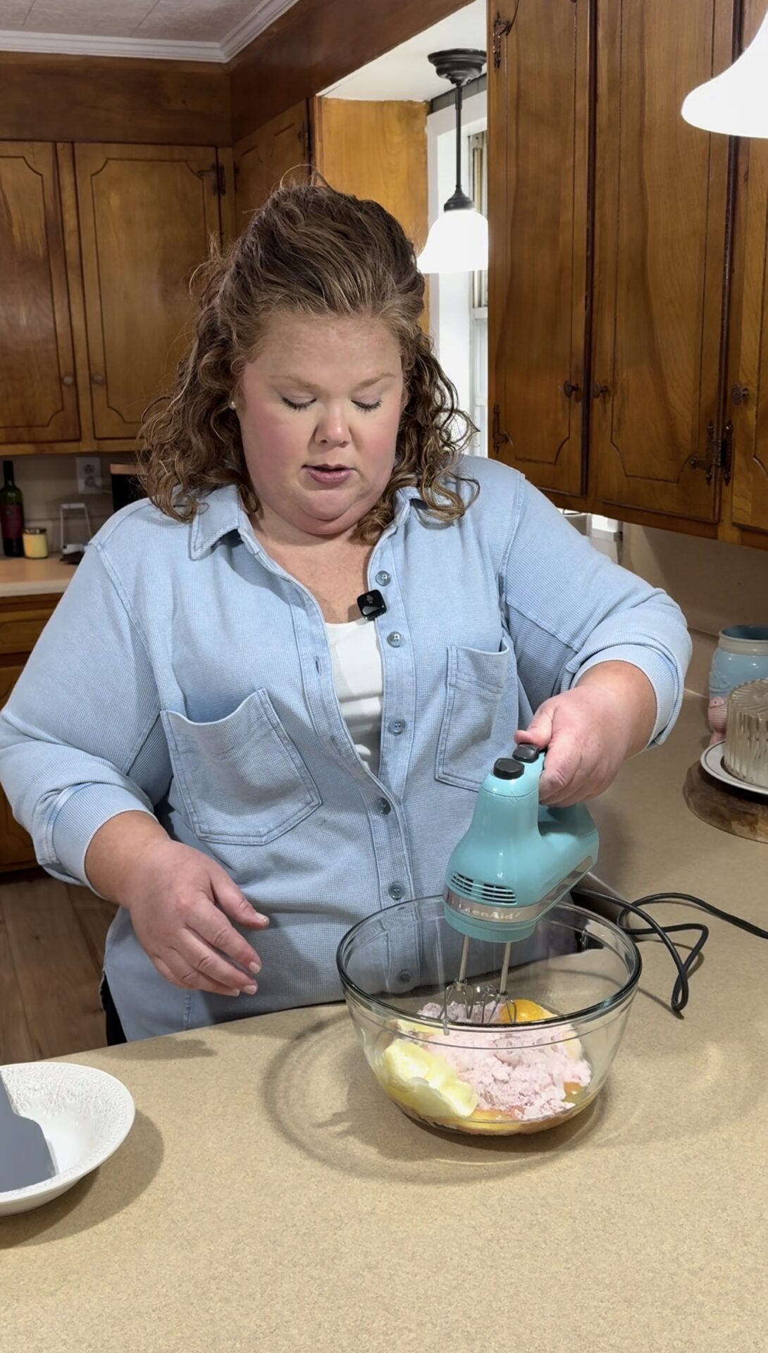 A woman in a light blue shirt uses a turquoise hand mixer to blend ingredients, including eggs, in a glass bowl on a kitchen counter. Wooden cabinets and kitchen items are visible in the background.