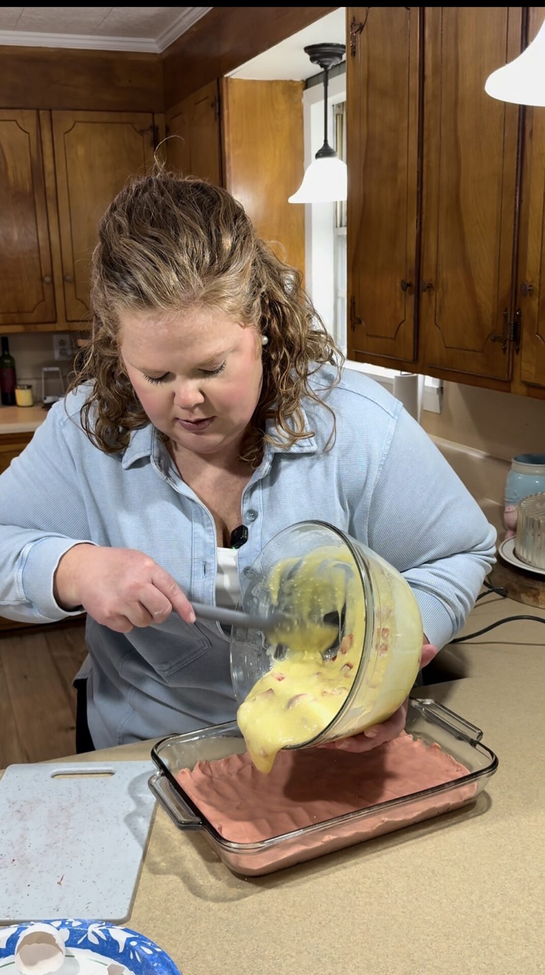 A woman in a light blue shirt pours yellow batter with fruit from a glass bowl into a baking dish with a pink layer, working in a cozy kitchen with wooden cabinets.