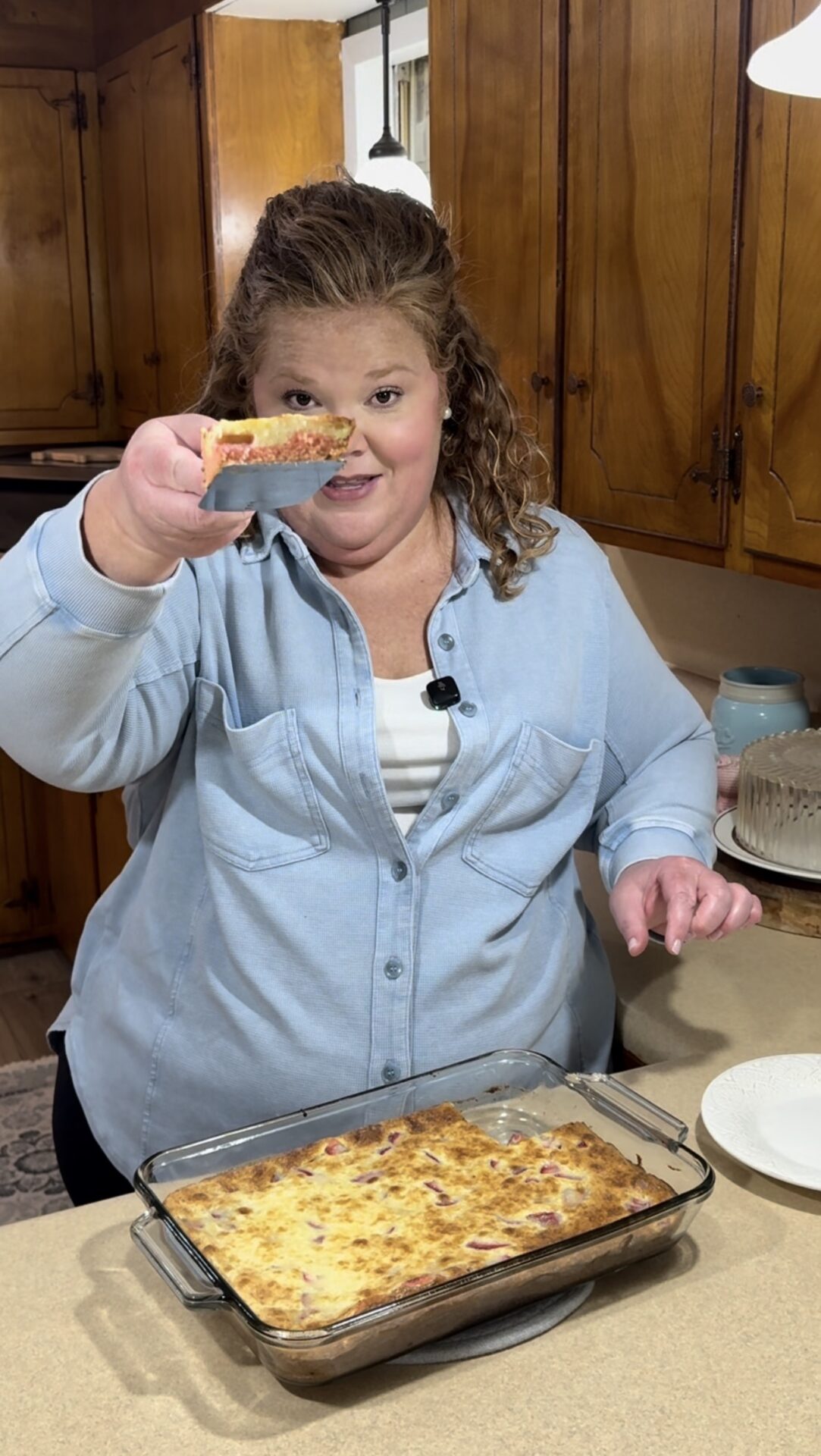 A woman in a light blue shirt holds up a slice of baked casserole in a kitchen, smiling at the camera. A glass baking dish with more casserole sits on the counter in front of her.