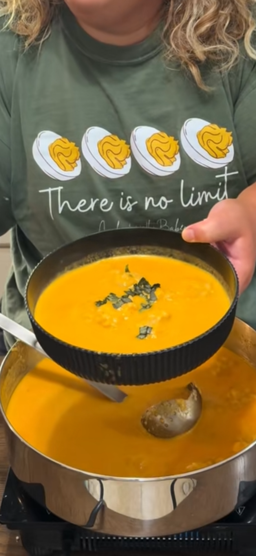 A person in a green shirt with egg illustrations holds a black bowl of orange soup over a pot, garnished with herbs, while serving it with a ladle.