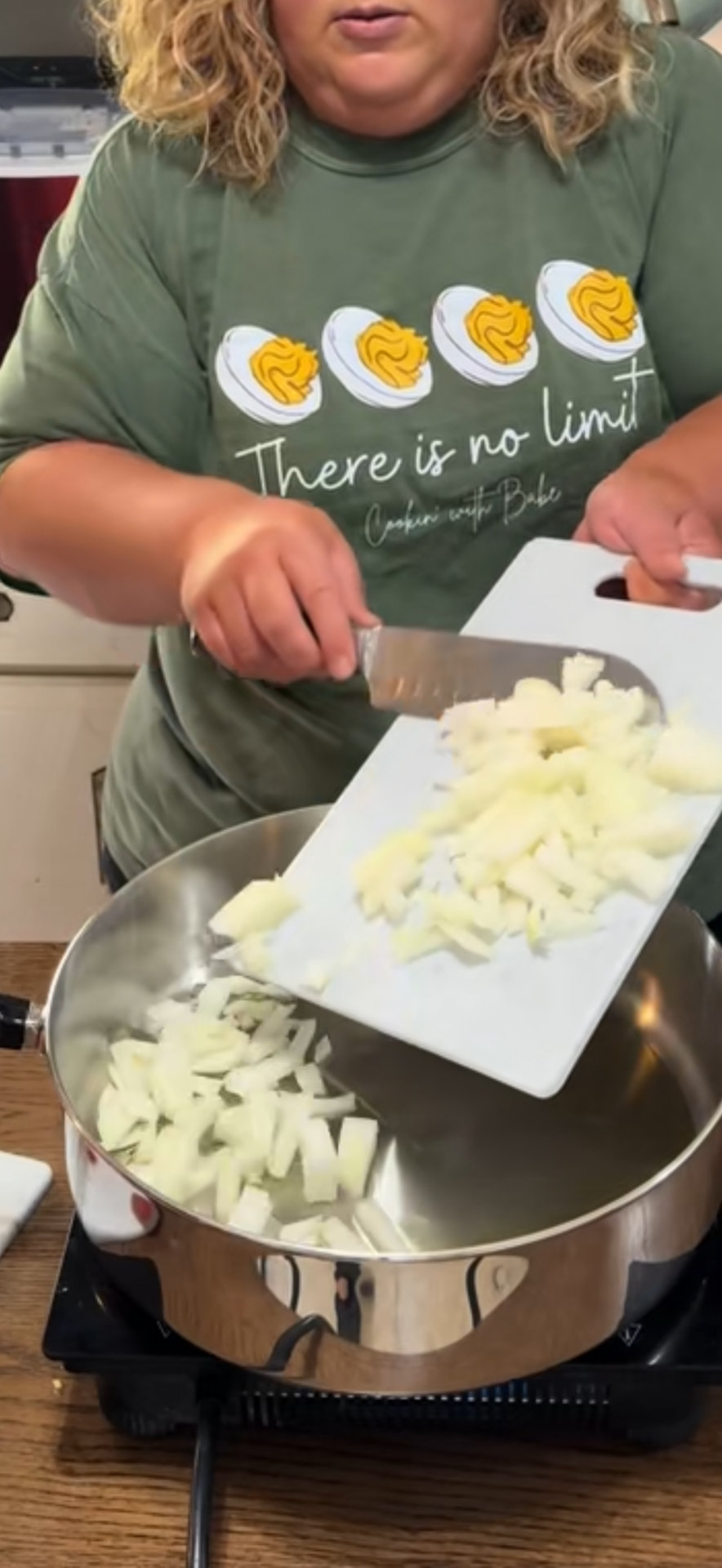 A person wearing a green shirt with images of eggs and the text “There is no limit” pours chopped onions from a cutting board into a stainless steel pan on a stove. Recipe is for Tomato Soup with Cheese Dumplings
