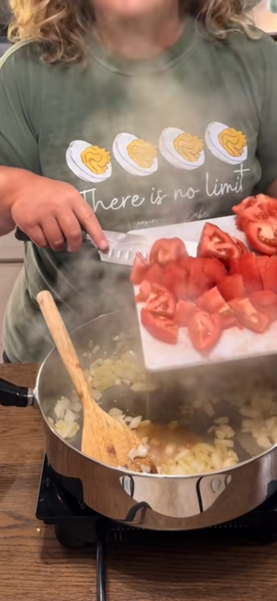 A person in a green shirt with eggs printed on it pours chopped tomatoes from a cutting board into a pot of onions cooking on a stove, as steam rises from the pot.
