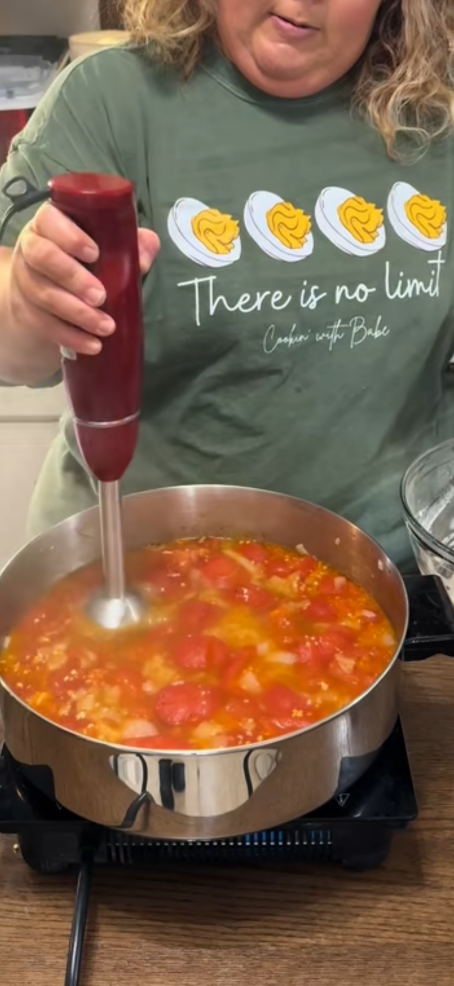 A woman using a red immersion blender to puree tomatoes and vegetables in a large pan on a stovetop. She is wearing a green shirt with cartoon eggs and the text There is no limit.