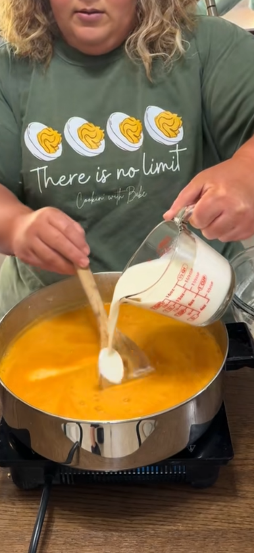 A person pours milk from a measuring cup into an orange soup while stirring with a wooden spoon. The person is wearing a green shirt with images of eggs and the text There is no limit.