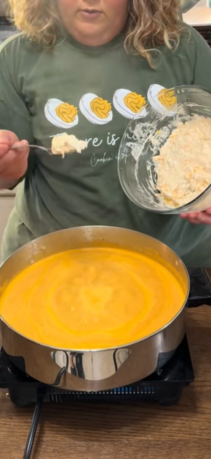 A person holds a spoonful of creamy mixture over a bowl in one hand and is about to add it to a pot of orange soup on a stovetop. The person is wearing a green shirt with deviled eggs printed on it.