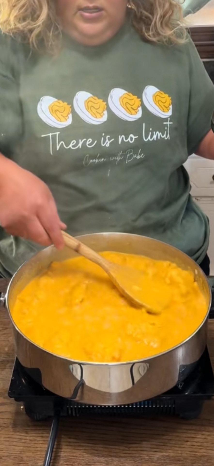 A person stirs a large pan of yellow, creamy food on a stove. They wear a green shirt with images of cut boiled eggs and the text There is no limit.