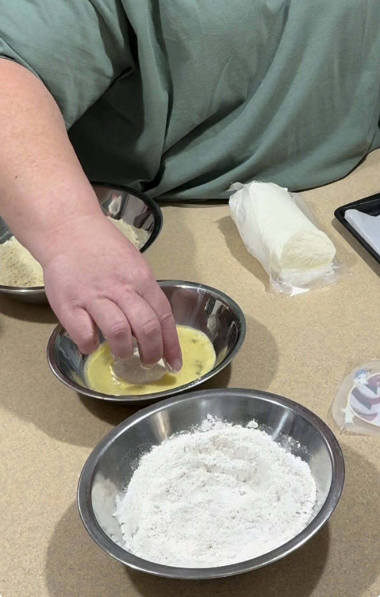 A person dips food into a bowl of beaten eggs, with bowls of flour and breadcrumbs nearby. A log of cheese and a tablet are also on the countertop.