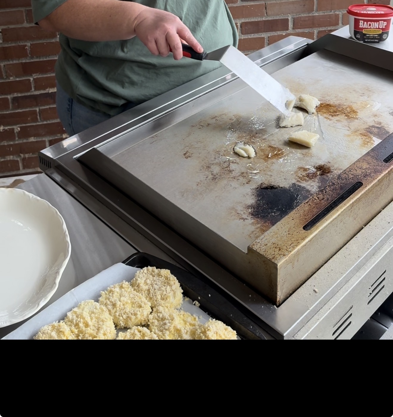 A person uses a spatula to cook food on a griddle, with some breaded items on a tray nearby and a tub of Bacon Up bacon grease on the side. The setting appears to be outdoors with a brick wall in the background.