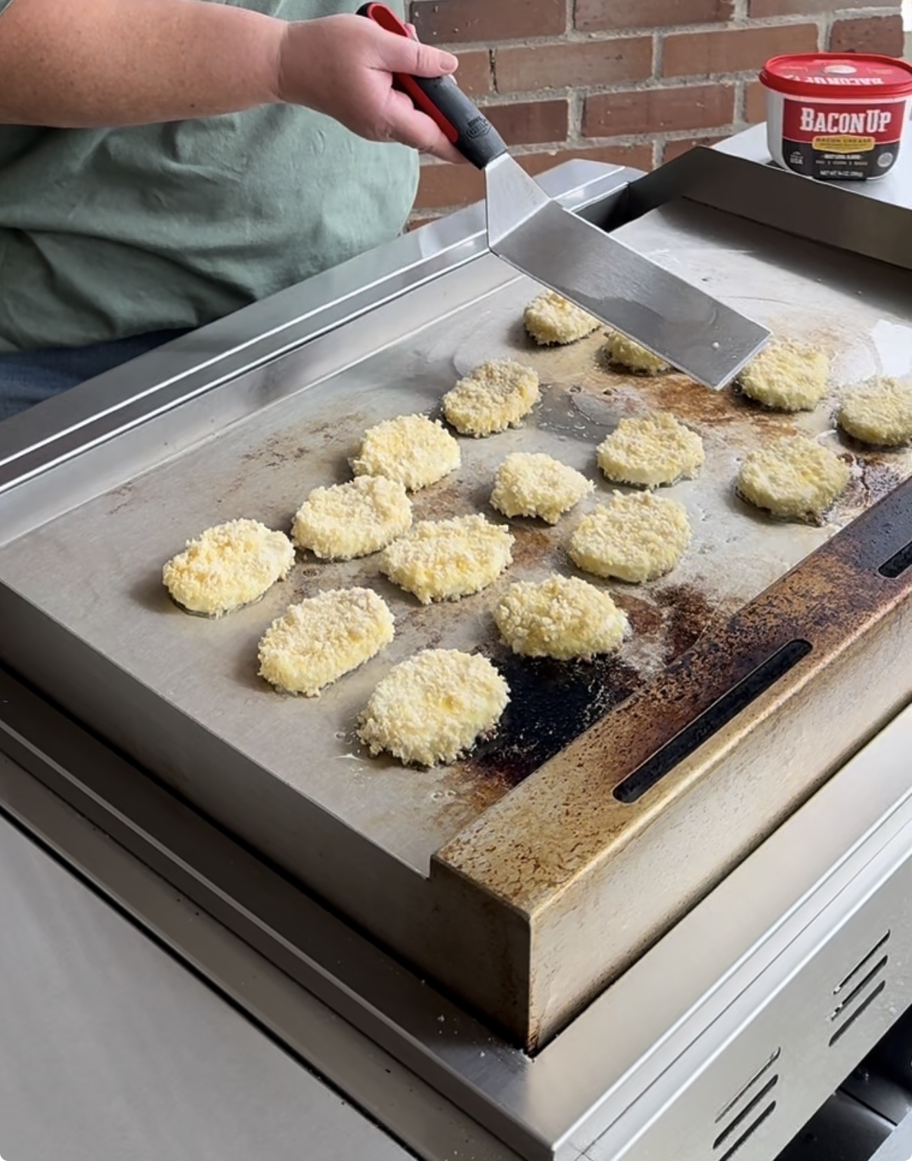 A person uses a metal spatula to cook breaded food pieces, possibly chicken or potato patties, on a large flat-top grill. A tub labeled Bacon Up is in the background near a brick wall.