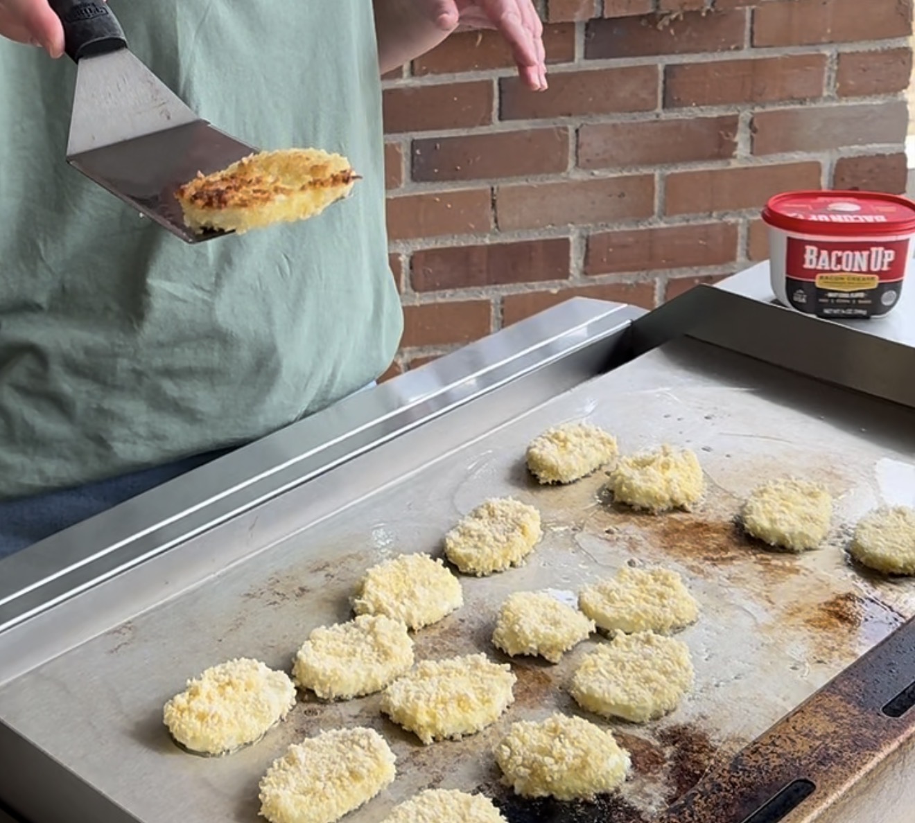 A person cooks breaded chicken nuggets on a flat-top grill, holding one piece with a spatula. A tub labeled Bacon Up is on the counter, and a brick wall is in the background.