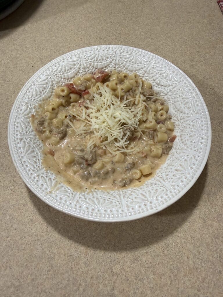 A white textured bowl filled with creamy pasta mixed with ground meat and tomatoes, topped with shredded cheese, sits on a beige countertop.