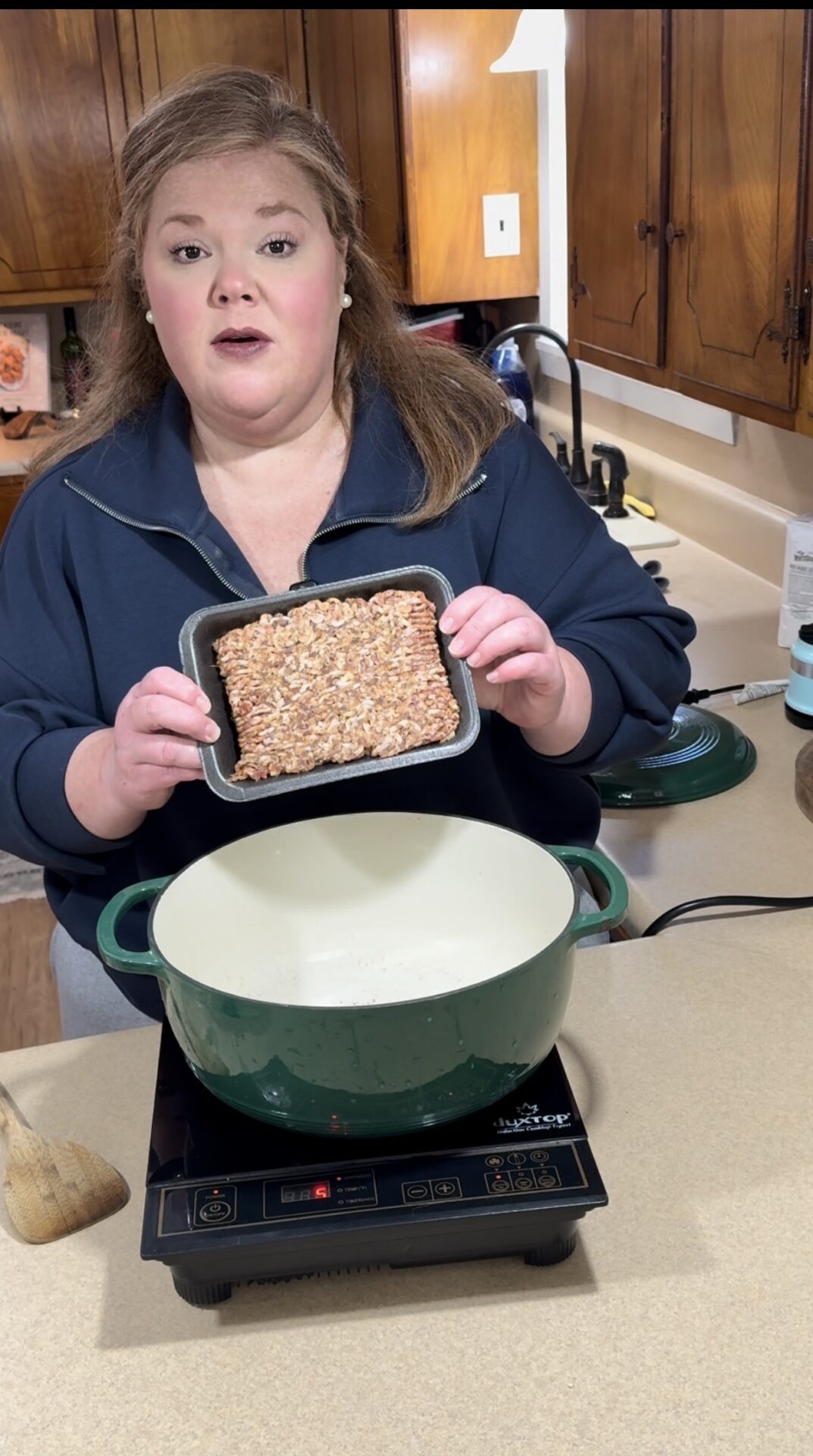 A woman stands in a kitchen holding a pan of uncooked ground meat over a large green pot on an induction cooktop, preparing to cook. Wooden cabinets and kitchen utensils are visible in the background.  Recipe for Creamy Italian Sausage Soup